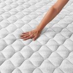 Suave y acolchada, vista cercana del colchón con mano tocando la superficie, en un entorno de mobiliario y decoración moderna, ideal para camas y dormitorios.