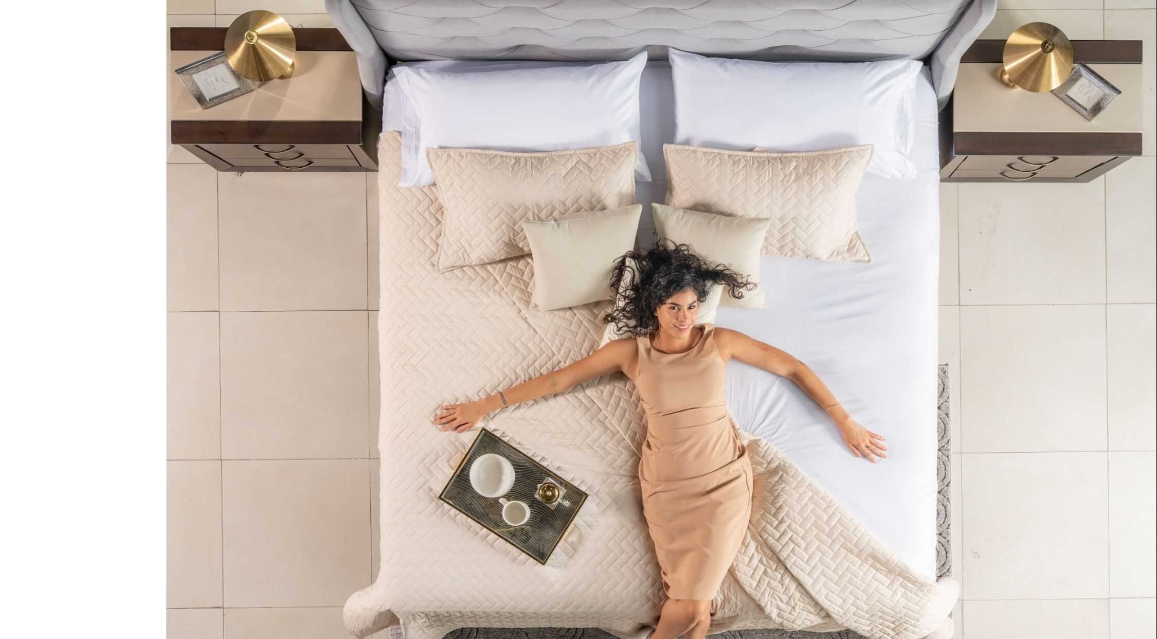 Suelo de baldosas blancas y cómoda cama con ropa de cama beige y múltiples cojines, con una mujer sonriendo en la cama, decoración moderna y elegante en la habitación.