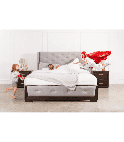 Muñeco de nieve vestido con traje rojo y capa saltando sobre la cama con niños jugando en un dormitorio decorado con muebles de madera oscura, una lámpara y paredes blancas.